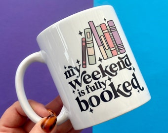 以下が含まれることがあります： 白いセラミック製のマグカップで、黒いデザインで「my weekend is fully booked」と書かれており、テキストの後ろにカラフルな本の積み重ねがあります。
