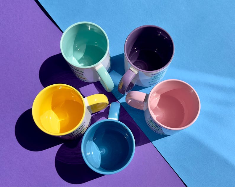 May include: Five colorful ceramic mugs with matching handles and interiors are arranged on a blue and purple surface. The mugs are in shades of yellow, blue, pink, purple, and teal. Each mug has a white exterior with text.