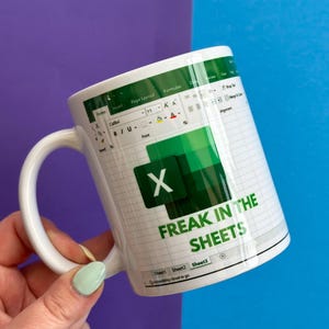 May include: White ceramic mug with a green Excel spreadsheet design, the Microsoft Excel logo, and the text "FREAK IN THE SHEETS". The mug has a white handle. The background is purple and blue.