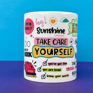 May include: White ceramic mug with colorful text and illustrations. The mug features phrases like "Sunshine," "Take Care of Yourself," and "You've Got This." Other graphics include a sun, clouds, and a stack of books. The mug is cylindrical.