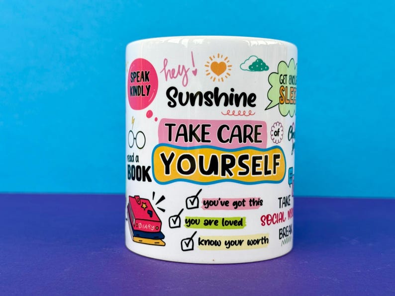 May include: White ceramic mug with colorful text and illustrations. The mug features phrases like "Sunshine," "Take Care of Yourself," and "You've Got This." Other graphics include a sun, clouds, and a stack of books. The mug is cylindrical.