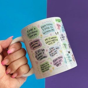 May include: White ceramic mug with a colorful design featuring positive affirmations and motivational quotes. The mug has a handle and is decorated with rainbows, flowers, and butterflies.