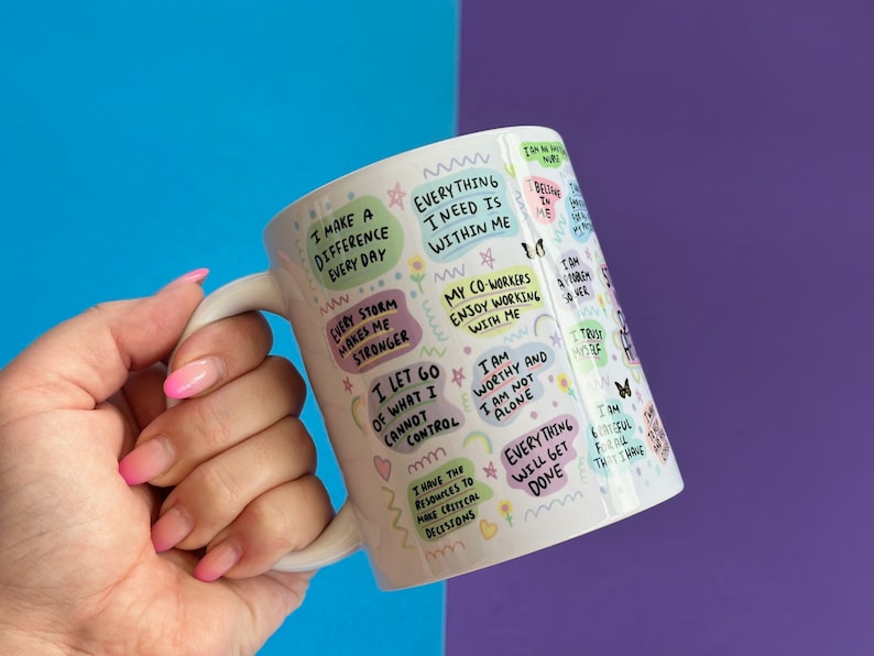 May include: White ceramic mug with a colorful design featuring positive affirmations and motivational quotes. The mug has a handle and is decorated with rainbows, flowers, and butterflies.
