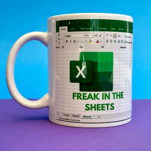 K&ouml;nnte beinhalten: Wei&szlig;e Keramiktasse mit einem Excel-Tabellen-Design und dem Text "FREAK IN THE SHEETS". Die Tasse hat einen wei&szlig;en Henkel und ein gr&uuml;n-wei&szlig;es Design. Die Tasse steht auf einer lila Oberfl&auml;che mit blauem Hintergrund.