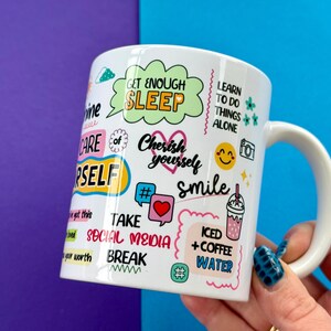 May include: White ceramic mug with a variety of colorful text and graphics, including "Get Enough Sleep," "Cherish Yourself," and "Smile." Other text includes "Take Social Media Break" and "Iced + Coffee Water."