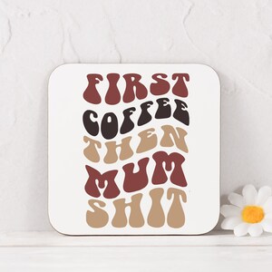 May include: A white coaster with the text "FIRST COFFEE THEN MUM SHIT" in a retro font with brown, red, and black colors.