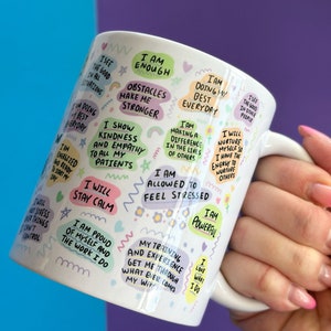 May include: White ceramic mug with a colorful design featuring positive affirmations and motivational quotes. The mug has a handle and is decorated with rainbows, stars, and flowers. The text on the mug includes phrases like "I am enough", "I will stay calm", and "I am allowed to feel stressed".