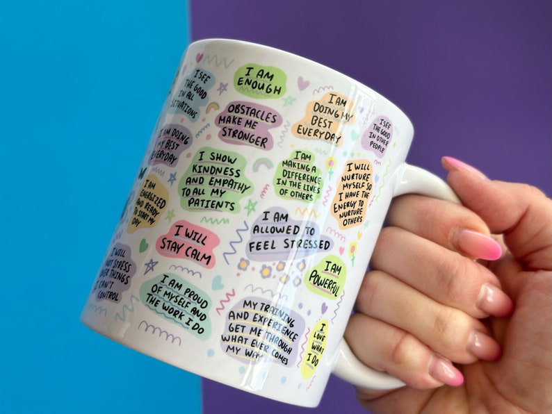 May include: White ceramic mug with a colorful design featuring positive affirmations and motivational quotes. The mug has a handle and is decorated with rainbows, stars, and flowers. The text on the mug includes phrases like "I am enough", "I will stay calm", and "I am allowed to feel stressed".
