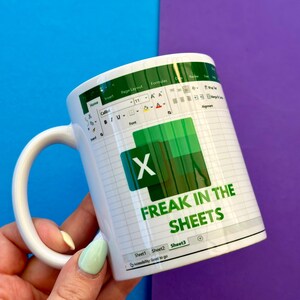 K&ouml;nnte beinhalten: Wei&szlig;e Keramik-Tasse mit einem gr&uuml;n-wei&szlig;en Excel-Tabellen-Design und dem Text "FREAK IN THE SHEETS". Die Tasse hat einen wei&szlig;en Henkel. Der Hintergrund ist blau und lila.