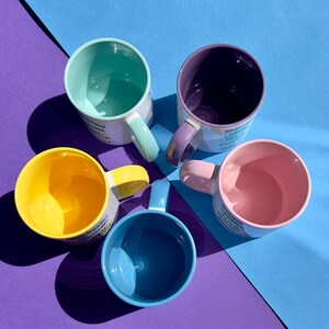 May include: Five ceramic mugs with different colored interiors and white exteriors. The mugs are arranged in a circle on a purple and blue background. Each mug has a different color interior: light blue, yellow, pink, purple, and green.
