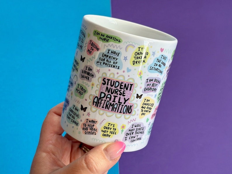 May include: White ceramic mug with colorful text and illustrations. The text reads "Student Nurse Daily Affirmations" and includes phrases like "I am an amazing nurse", "I have empathy for all of my patients", "I am a problem solver", "I trust myself", "I am grateful for all that I have", "I want to help and heal others", "It's okay to not feel okay", "I will not stress over things I can't control", "I see the good in all situations", "I am doing my best everyday", "I am energized and ready to start my day".