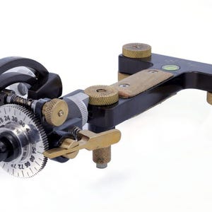 May include: A precision instrument with a black body, brass accents, and a silver dial featuring numbered markings. The device has a needle-like tip and adjustable knobs, suggesting it's used for detailed work or measurement.