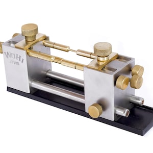 May include: A silver-colored metal tool with brass knobs and rods. The tool has the text "WOHI JTWS" engraved on the side. The tool sits on a black base and has multiple adjustable parts.