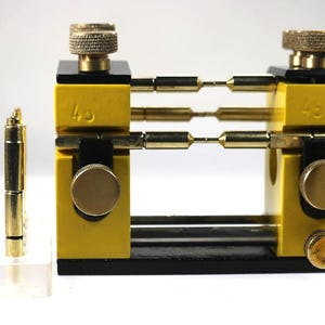 Puede incluir: Un conjunto de herramientas de relojería, que incluye un soporte para el movimiento del reloj amarillo y negro con componentes de latón, y una herramienta de nivelación amarilla con el texto "WOHI". También se incluyen varias piezas de reloj doradas.