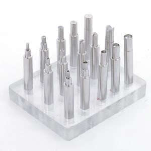 Puede incluir: Colección de varillas metálicas plateadas de diversas formas y tamaños, dispuestas sobre un expositor rectangular transparente. Las varillas presentan diseños cilíndricos, escalonados y ranurados, sobre un fondo blanco.