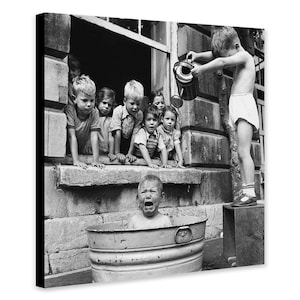 Op de afbeelding: Zwart-witfoto van spelende kinderen. Een kind giet water uit een gieter op een ander kind in een metalen teil. Andere kinderen kijken toe vanuit een raam.