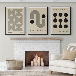 May include: Three framed prints with black and white geometric designs on a beige background. The prints feature the text "Bauhaus" and dates. The prints are hanging above a white brick fireplace.