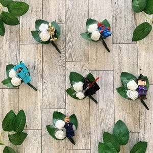 Pode incluir: Seis boutonnières de flores brancas com diferentes minifiguras Lego anexadas. As minifiguras incluem Spider-Man, Darth Vader, Capitão América, uma figura azul, uma figura com um capacete amarelo e uma figura com uma túnica roxa.