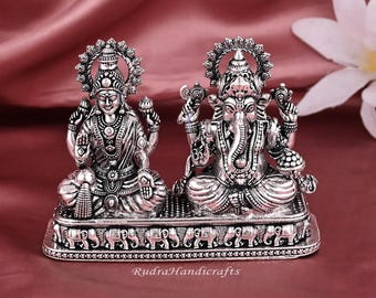 Può includere: Una statua color argento raffigurante le divinità Lakshmi e Ganesha, sedute fianco a fianco su una base decorata con motivi di elefanti. Le statue dettagliate sono poste su uno sfondo bordeaux. Il testo "RudraHandicrafts" è visibile sulla base.
