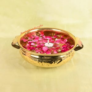 Könnte beinhalten: Eine goldfarbene Messingschale gefüllt mit Wasser, rosa Rosenblättern und einer brennenden weißen Kerze. Die Schale hat dekorative Griffe und ein geprägtes Design am Boden. Der Text "Rudra Handicrafts" ist sichtbar.