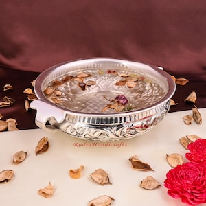 Könnte beinhalten: Eine silberne Schale mit aufwendigen floralen Designs, gefüllt mit Wasser und getrockneten Blumen. Die Schale hat zwei Griffe und steht auf einer weißen Oberfläche mit verstreuten getrockneten Blütenblättern.