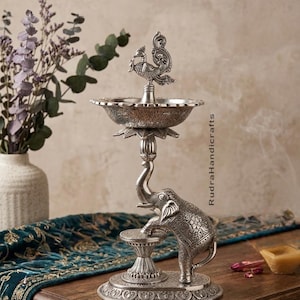 Op de afbeelding: Een zilverkleurige decoratieve lamp met een olifant- en pauwontwerp. De lamp heeft een gedetailleerde olifant die een klein platform ondersteunt, met een pauw boven een komvormig reservoir. De lamp staat op een houten oppervlak.
