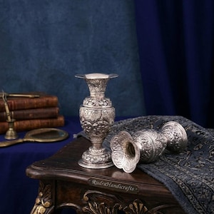 Può includere: Vasi d'argento ornati con dettagliati motivi floreali. Un vaso è in posizione verticale, mentre altri due sono sdraiati. Sono su un tavolo di legno scuro con uno sfondo blu. Il testo "RudraHandicrafts" è visibile sul tavolo.