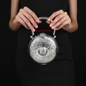 May include: A silver round clutch purse with an ornate floral design and a silver handle. The purse is held by a person's hands.