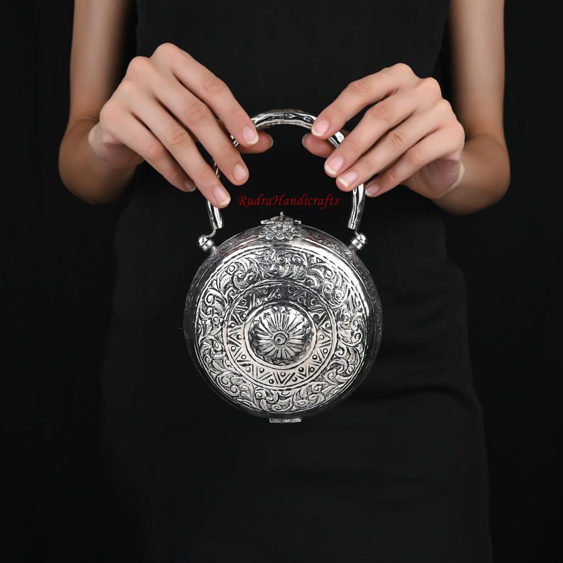 May include: A silver round clutch purse with an ornate floral design and a silver handle. The purse is held by a person's hands.