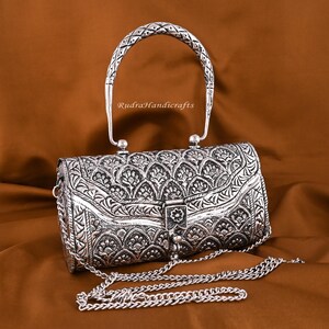 May include: Silver-colored, ornate handbag with a patterned design and a chain strap. The handbag has a curved handle and a clasp closure. The text "RudraHandicrafts" is visible on the handle.