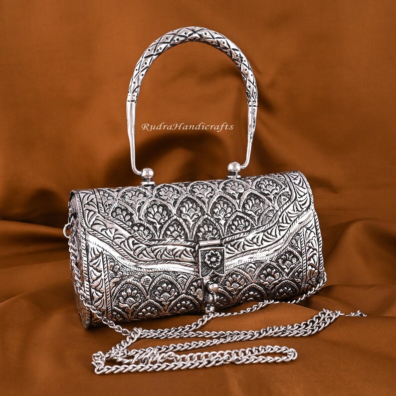 May include: Silver-colored, ornate handbag with a patterned design and a chain strap. The handbag has a curved handle and a clasp closure. The text "RudraHandicrafts" is visible on the handle.