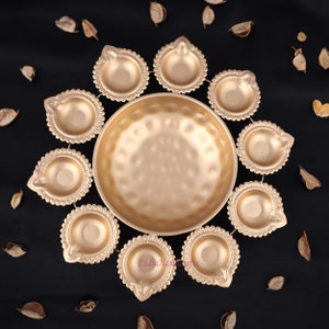 以下が含まれることがあります： 金色の装飾用オイルランプと中央のボウルのセットで、円形に配置されています。 ランプはスカラップエッジのデザインで、散らばった乾燥した葉のある暗い背景に配置されています。 ボウルはハンマー加工されたテクスチャです。