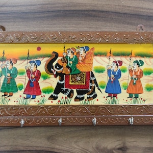 Może przedstawiać: Drewniany wieszak na klucze z namalowanym obrazem ludzi jadących na słoniu. Scena jest oprawiona w brązową drewnianą ramę z misternymi rzeźbami. Na dole wieszaka znajdują się pięć haczyków na klucze.