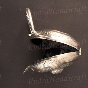 May include: A silver-colored, ornate, hinged box with an engraved design. The box is open, revealing a black velvet interior.