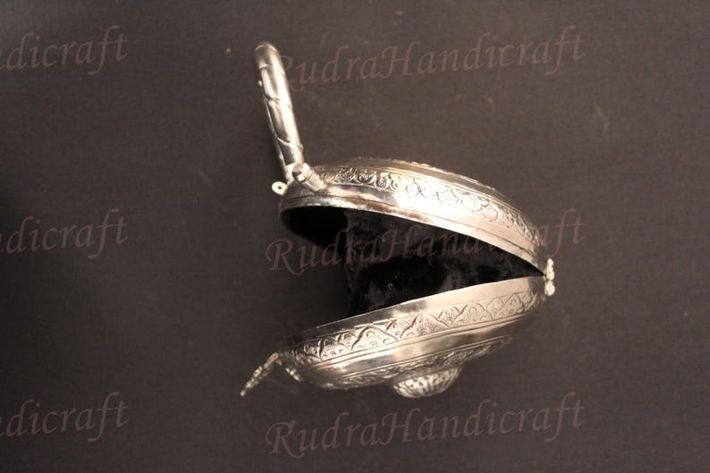 May include: A silver-colored, ornate, hinged box with an engraved design. The box is open, revealing a black velvet interior.