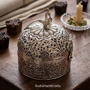 May include: A silver-toned metal container with a domed, perforated lid featuring intricate floral engravings and a bird-shaped handle. The cylindrical base displays detailed embossed leaf patterns. The text RudraHandicrafts is visible.
