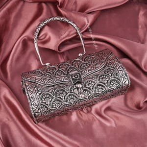 May include: Silver-colored, intricately carved handbag with a curved handle. The bag features a floral pattern and a clasp closure. The handle is braided. The bag is set against a pink satin background.