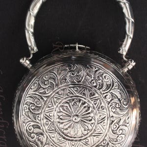 May include: A silver round box with an ornate floral design and a silver handle. The box has a hinged lid and a small latch.