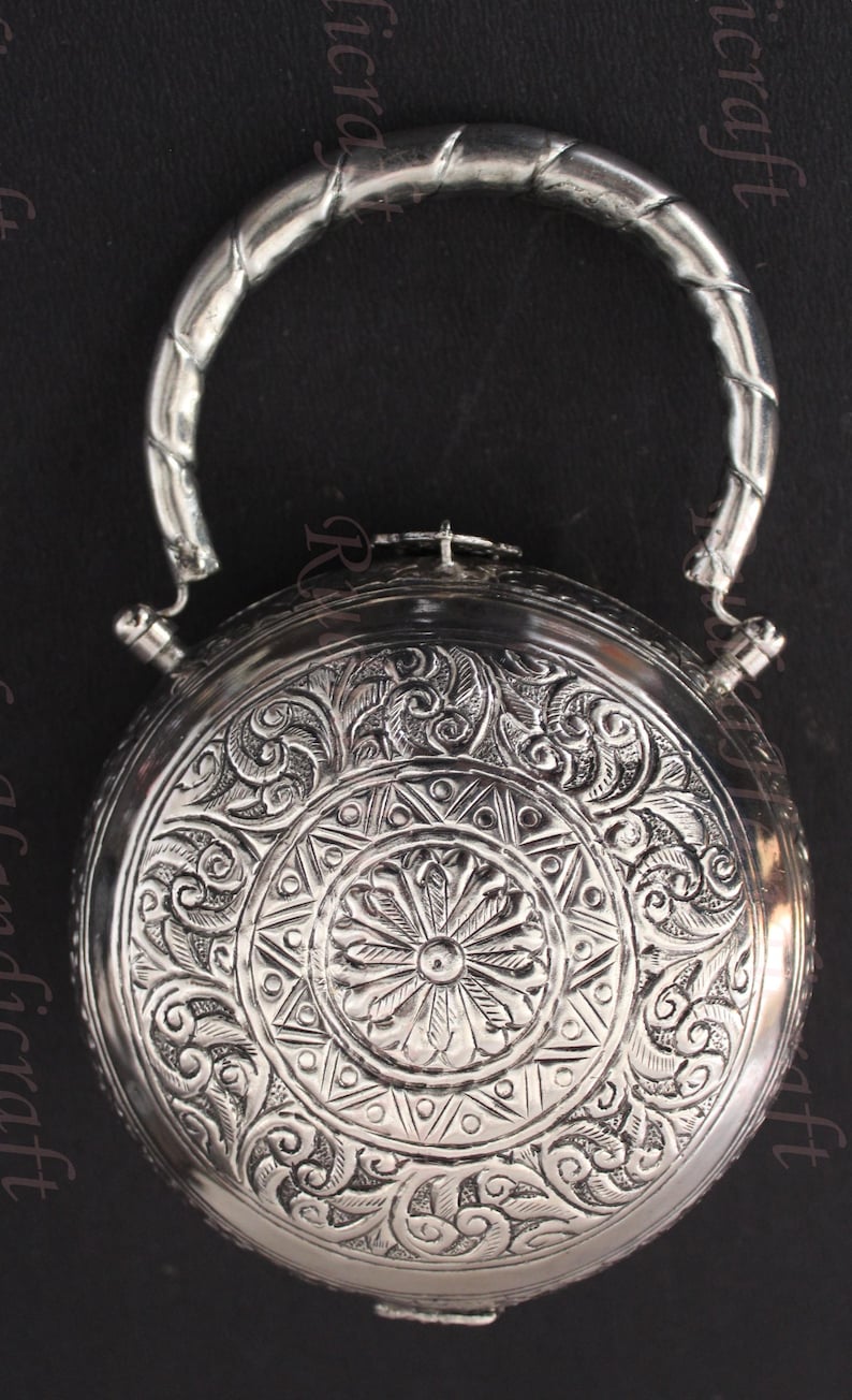 May include: A silver round box with an ornate floral design and a silver handle. The box has a hinged lid and a small latch.