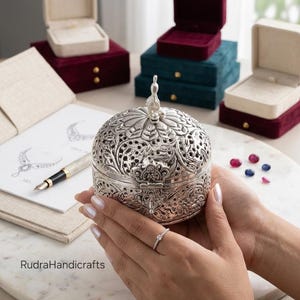 May include: A round, silver-toned metal box featuring intricate floral filigree cutouts and a decorative finial on the lid. The box is held in place, with a fountain pen, a sketch of jewelry, and several velvet jewelry boxes visible nearby.