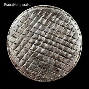 May include: A circular silver-toned object with a textured grid pattern of small raised squares across its surface. The text RudraHandicrafts is visible in the upper left corner, and the text BOTTOM SIDE is printed at the bottom of the image.