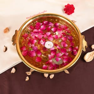 以下が含まれることがあります： 水とピンクのバラの花びらで満たされた金色の金属製のボウル。小さな白いろうそくが中央に浮かんでいます。ボウルには装飾的なハンドルがあり、乾燥した花びらと赤い花に囲まれています。