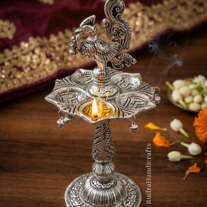 May include: A silver-colored, ornate oil lamp with a peacock design on top. The lamp has a round base and a floral-shaped oil reservoir with a lit flame. Small bells hang from the reservoir. The background includes flowers and a maroon cloth.
