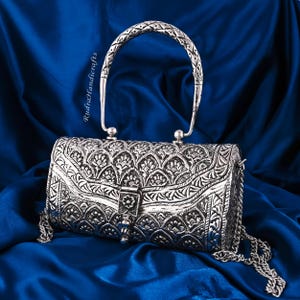 May include: A silver-colored, cylindrical handbag with intricate floral patterns and a chain strap. The bag features a decorative clasp and a textured handle. The background is a deep blue fabric.