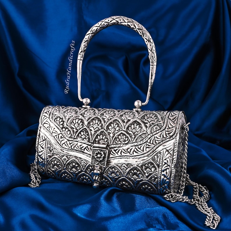 May include: A silver-colored, cylindrical handbag with intricate floral patterns and a chain strap. The bag features a decorative clasp and a textured handle. The background is a deep blue fabric.