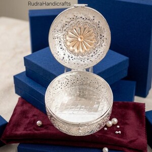 May include: A round silver-toned metal box with an open hinged lid, featuring intricate filigree floral patterns and a central sunburst design. The interior base has a grid-like texture. The text RudraHandicrafts is visible in the upper left.