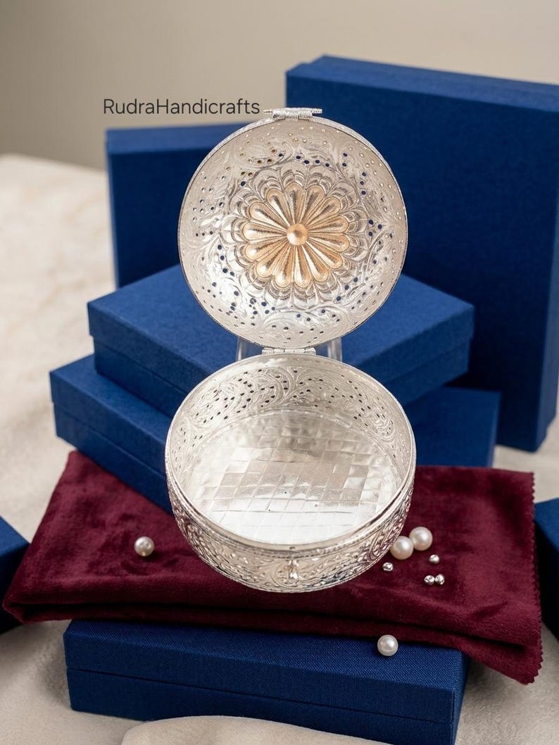 May include: A round silver-toned metal box with an open hinged lid, featuring intricate filigree floral patterns and a central sunburst design. The interior base has a grid-like texture. The text RudraHandicrafts is visible in the upper left.