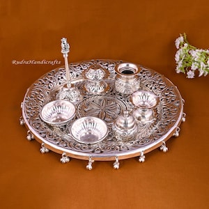 May include: A silver puja thali with intricate designs and a bell border. The thali includes a small silver diya, a silver incense burner, a silver water pot, a silver bell, and two silver bowls.