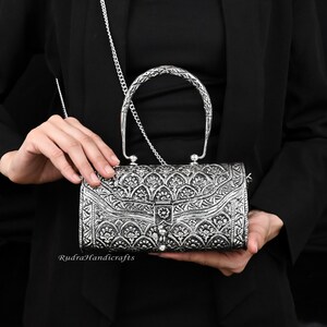 May include: Silver-colored rectangular handbag with intricate floral patterns and a braided handle. The bag has a chain strap and a clasp closure. The brand name "Rudra Handicrafts" is visible on the bottom left.
