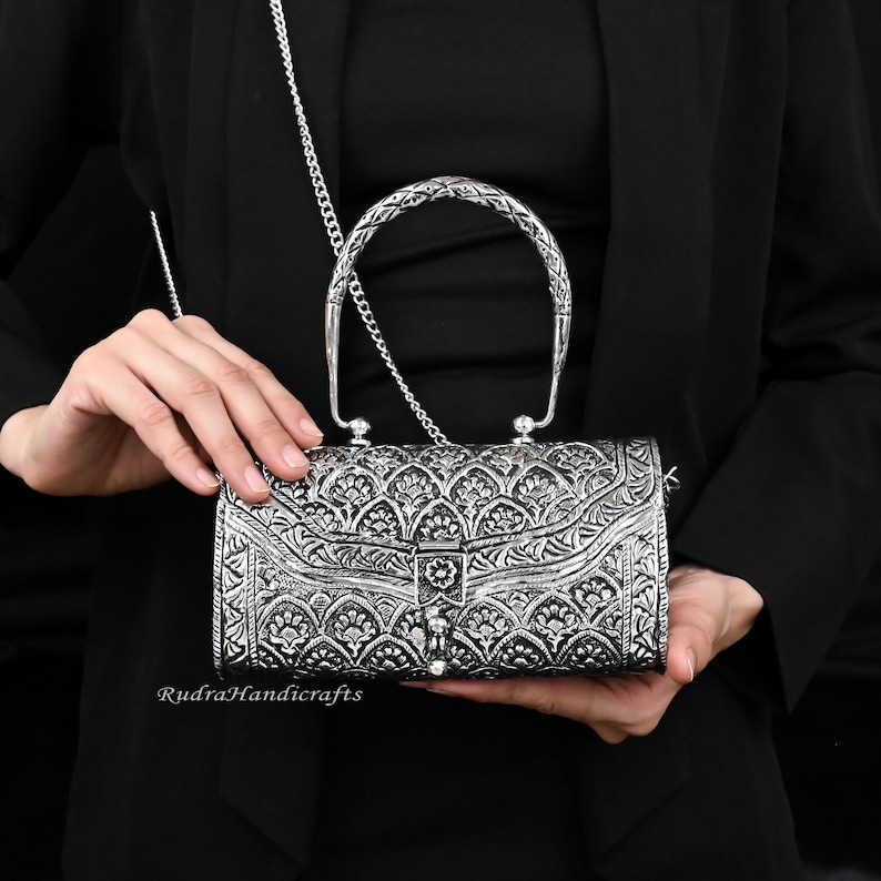 May include: Silver-colored rectangular handbag with intricate floral patterns and a braided handle. The bag has a chain strap and a clasp closure. The brand name "Rudra Handicrafts" is visible on the bottom left.